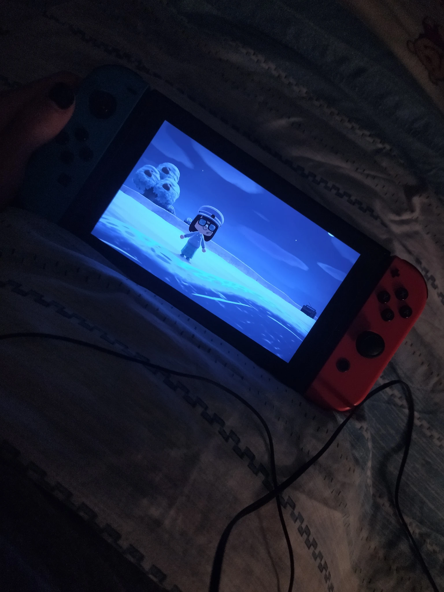 Foto alla mia console tenuta in mano mentre sono a letto, luce soffusa dell'ambiente che viene giusto dal lato dove ho la lampada, schermata molto blu del gioco a causa della sera, dove sono a riva della spiaggia per fare scena