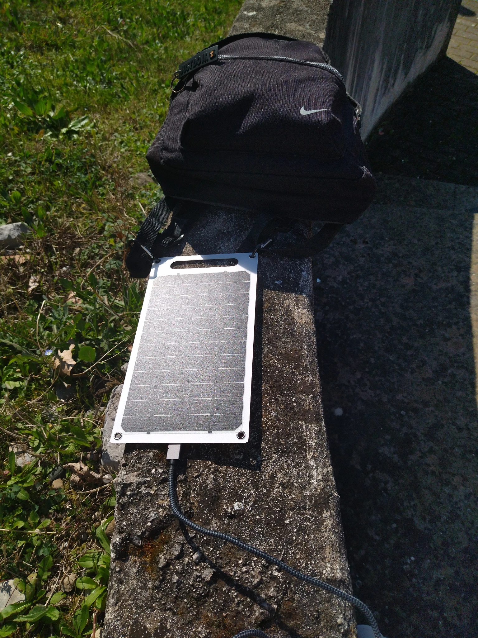 Foto al muretto su cui ero seduta, pannello solare in vista con sole battente e cavo USB collegato, attaccato allo zaino appoggiato affianco.